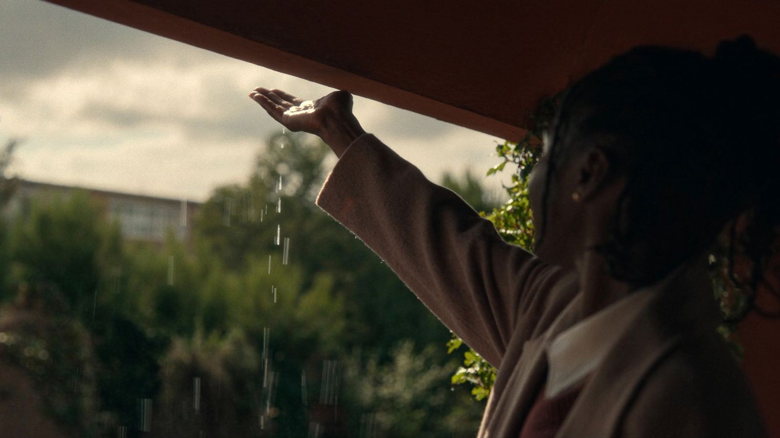 Iemand steekt vanonder een overkapping een hand uit in de regen, terwijl er verderop al zon doorbreekt